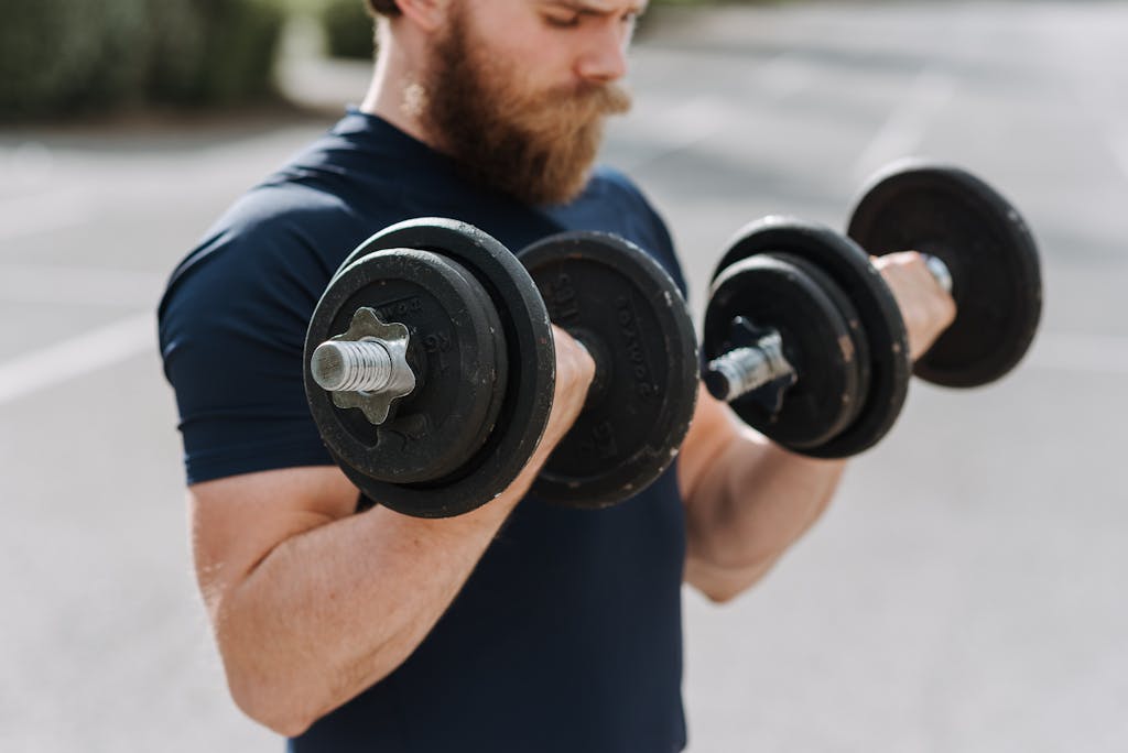 Crop sporty male making effort doing exercise with dumbbells while working out on blurred background of sports ground