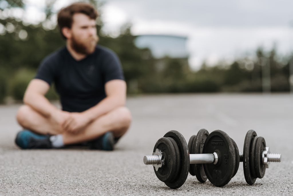 Young male with beard resting after workout on asphalt road with heavy dumbbells on gloomy day in selective focus