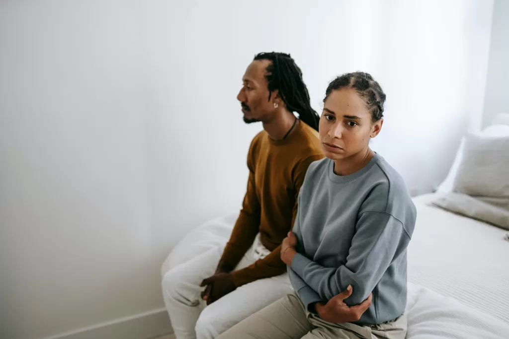 A couple sitting silently on a bed with tense expressions, suggesting conflict and emotional struggles.