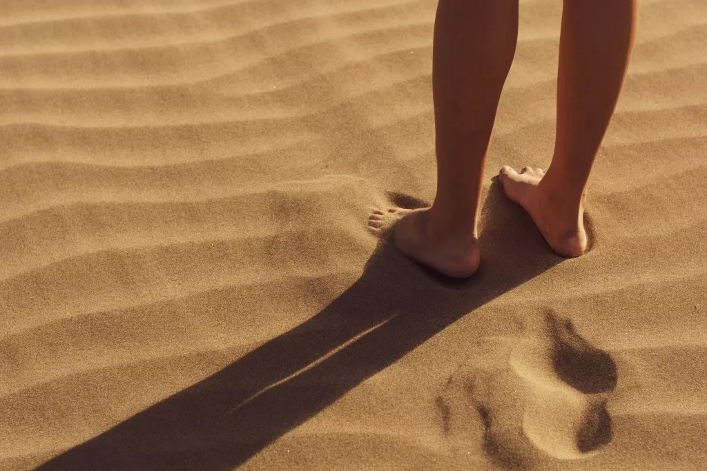 Close-up of a person standing barefoot on sunlit sand with visible footprints.