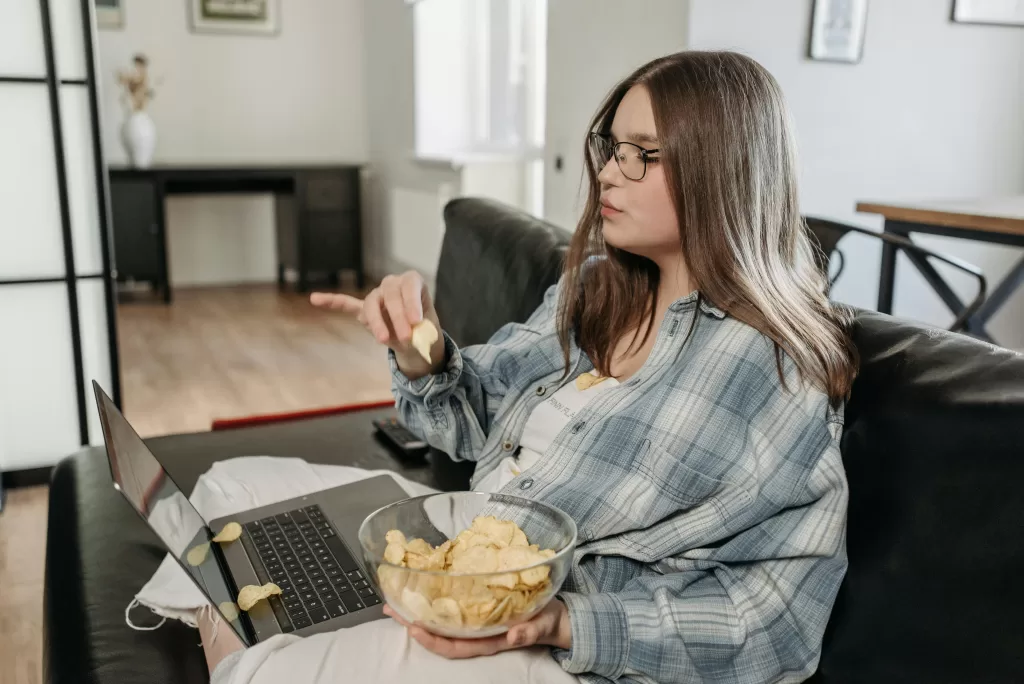 Woman in casual attire relaxing on a couch with a laptop and bowl of chips.